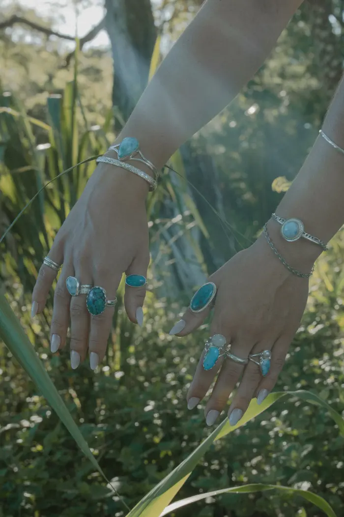 Two hands stacked with turquoise rings and silver cuffs among tropical leaves